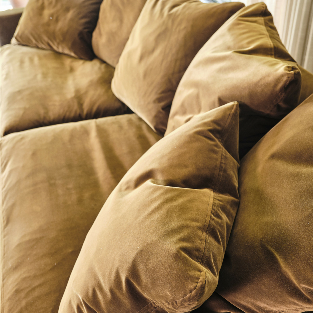 Un canapé en velours marron avec des coussins en peluche assortis, placé près d'une fenêtre avec de la verdure à l'extérieur. Canape Paris by Atelier Bis