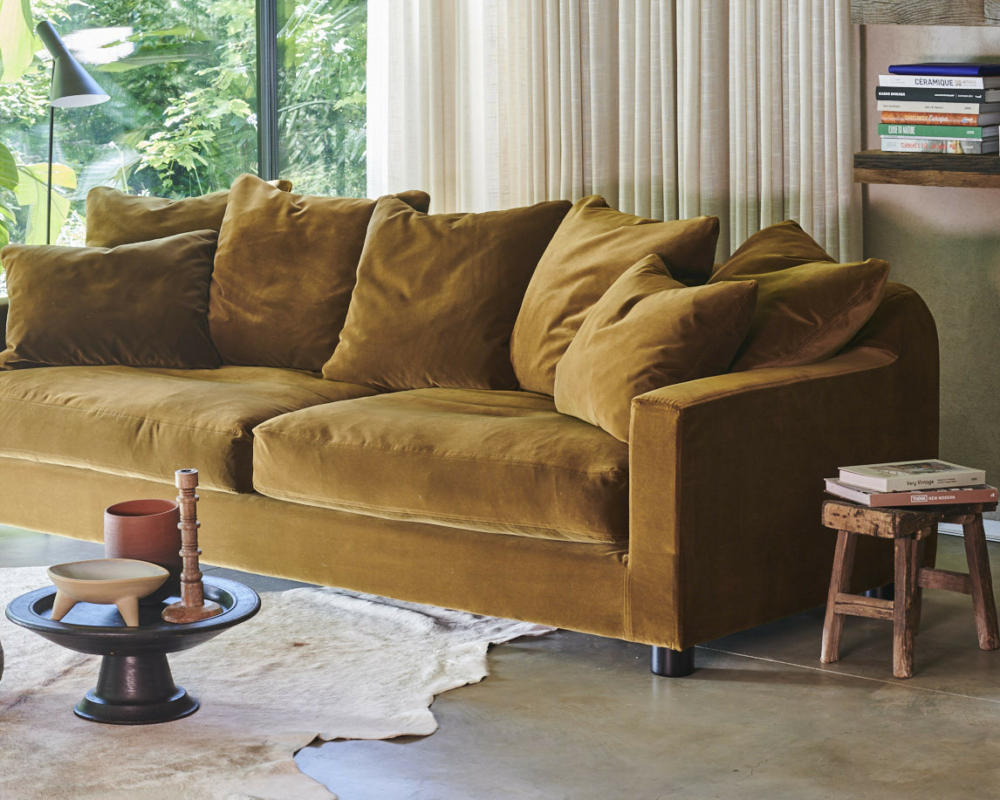 Un canapé en velours jaune moutarde avec des coussins moelleux est placé près d'une fenêtre, à côté de livres et de décorations en bois dans une pièce confortable. Canape Paris by Atelier Bis