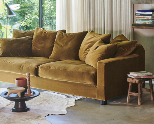 Un canapé en velours jaune moutarde avec des coussins moelleux est placé près d'une fenêtre, à côté de livres et de décorations en bois dans une pièce confortable. Canape Paris by Atelier Bis