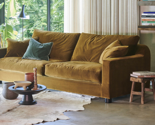 Un canapé en velours jaune moutarde avec des coussins est posé près d'une fenêtre dans un salon confortable et ensoleillé. Canape Paris by Atelier Bis