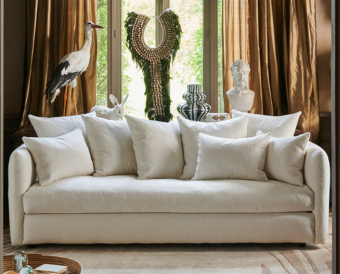 Canapé blanc avec de nombreux coussins devant une fenêtre, entouré de statues, d'un vase et de sculptures d'animaux. Canape Paris by Atelier Bis