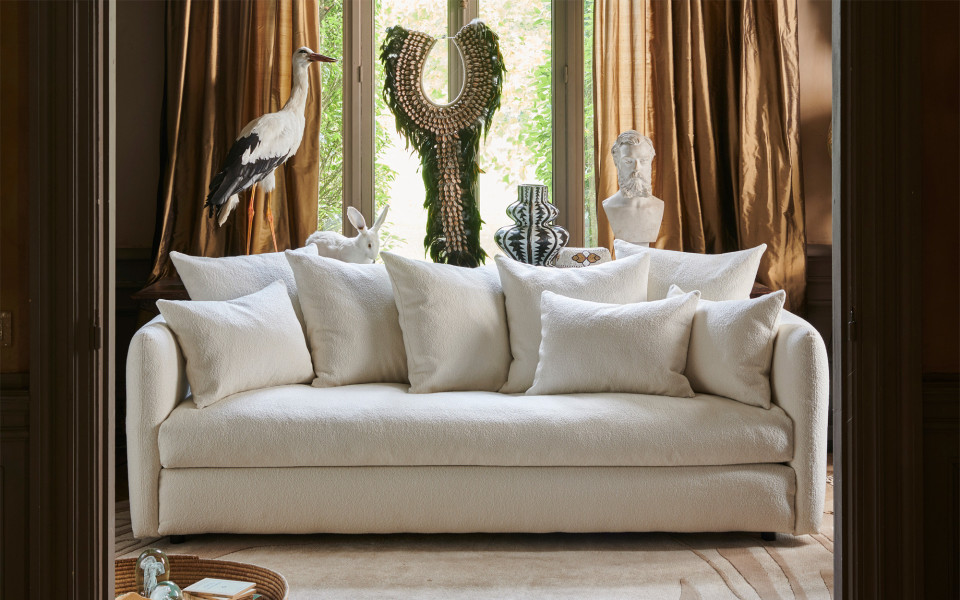 Canapé blanc avec de nombreux coussins devant une fenêtre, entouré de statues, d'un vase et de sculptures d'animaux. Canape Paris by Atelier Bis