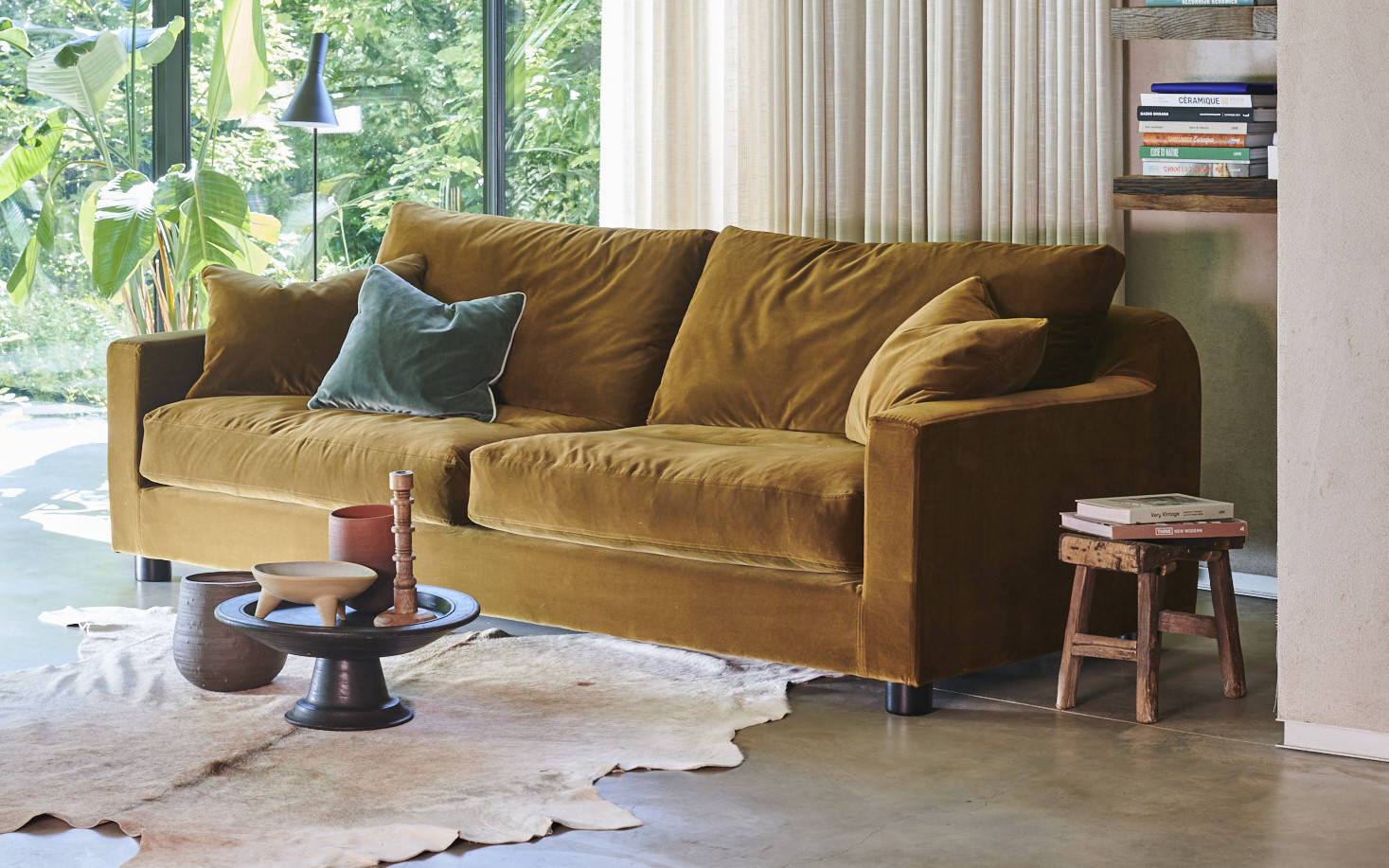 Un canapé en velours jaune moutarde avec des coussins est posé près d'une fenêtre dans un salon confortable et ensoleillé. Canape Paris by Atelier Bis