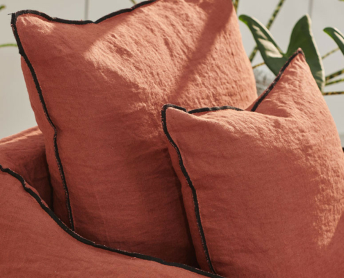 Deux coussins couleur rouille avec un passepoil noir sur une chaise assortie, avec des feuilles de plantes vertes en arrière-plan. Canape Paris by Atelier Bis