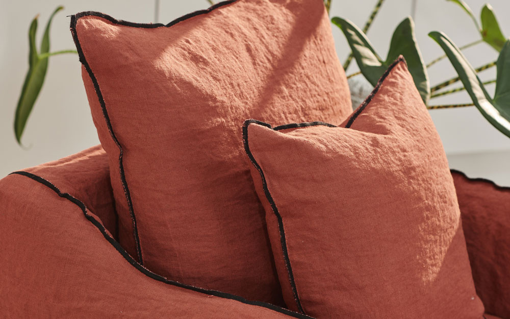 Deux coussins couleur rouille avec un passepoil noir sur une chaise assortie, avec des feuilles de plantes vertes en arrière-plan. Canape Paris by Atelier Bis