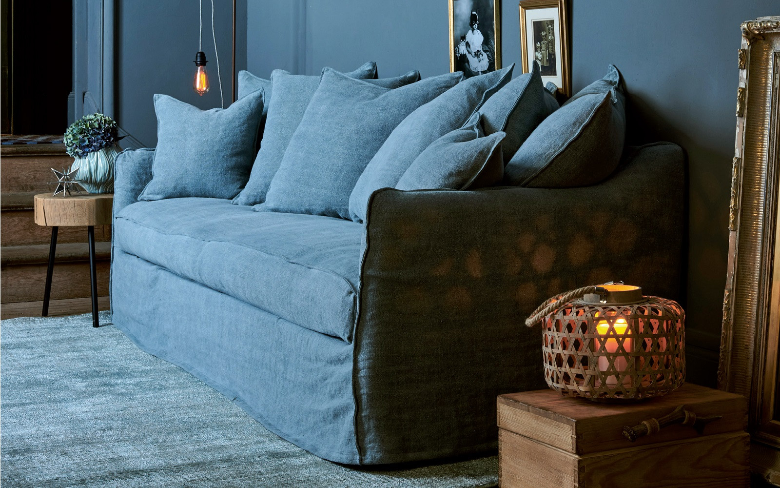 Canapé bleu avec de nombreux coussins dans un salon confortable avec un éclairage tamisé et des accents décoratifs sur des surfaces en bois. Canape Paris by Atelier Bis