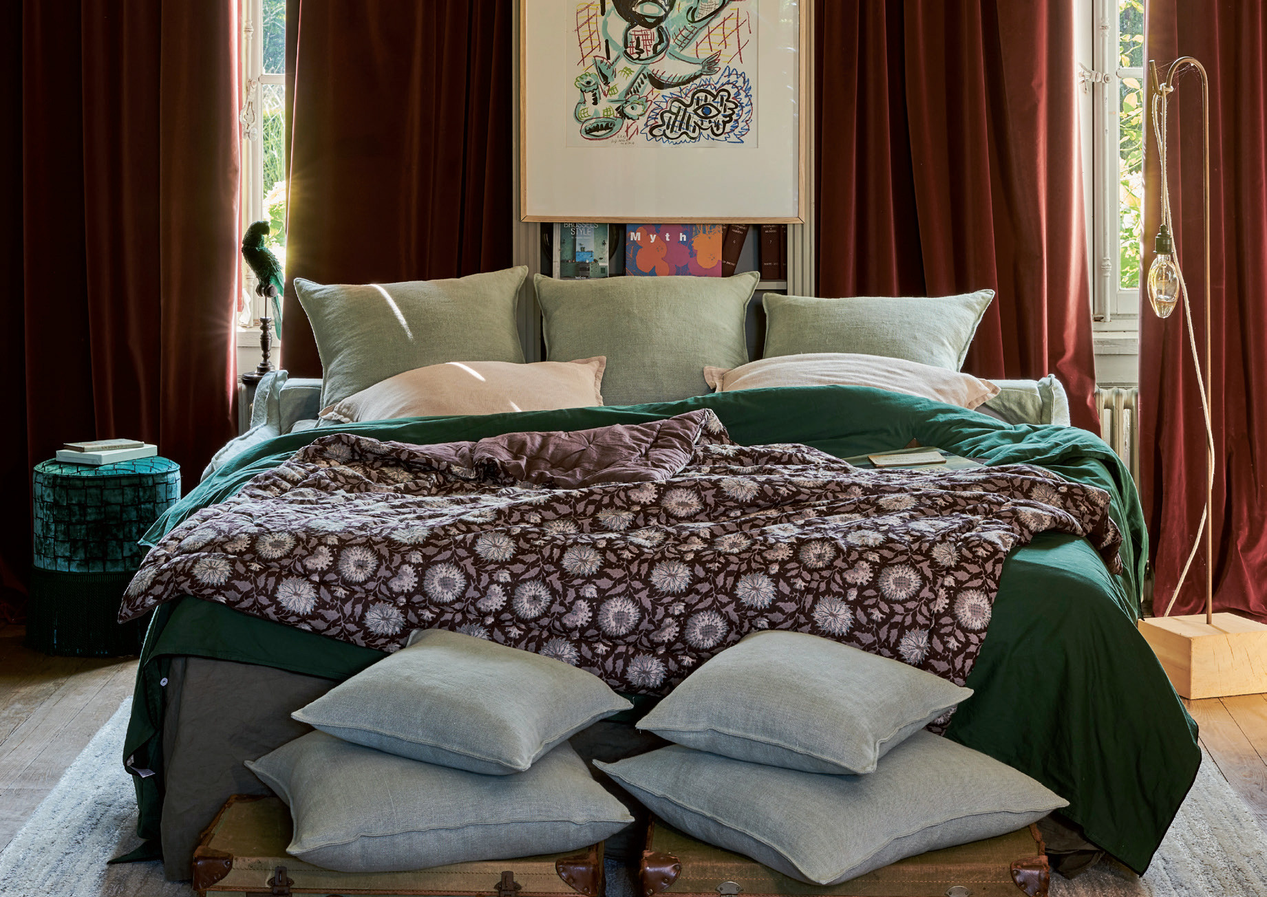 Un lit convertible Homesprit, confortable et sans canapé, avec une literie verte et brune, de nombreux oreillers et des œuvres d'art au-dessus, encadré par des rideaux bordeaux. Canape Paris by Atelier Bis