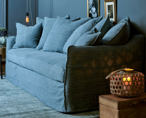 Canapé bleu convertible Homesprit Cassis avec de nombreux coussins dans une pièce confortable et peu éclairée, accentuée par une lanterne à bougie et des photos encadrées au mur. Canape Paris by Atelier Bis