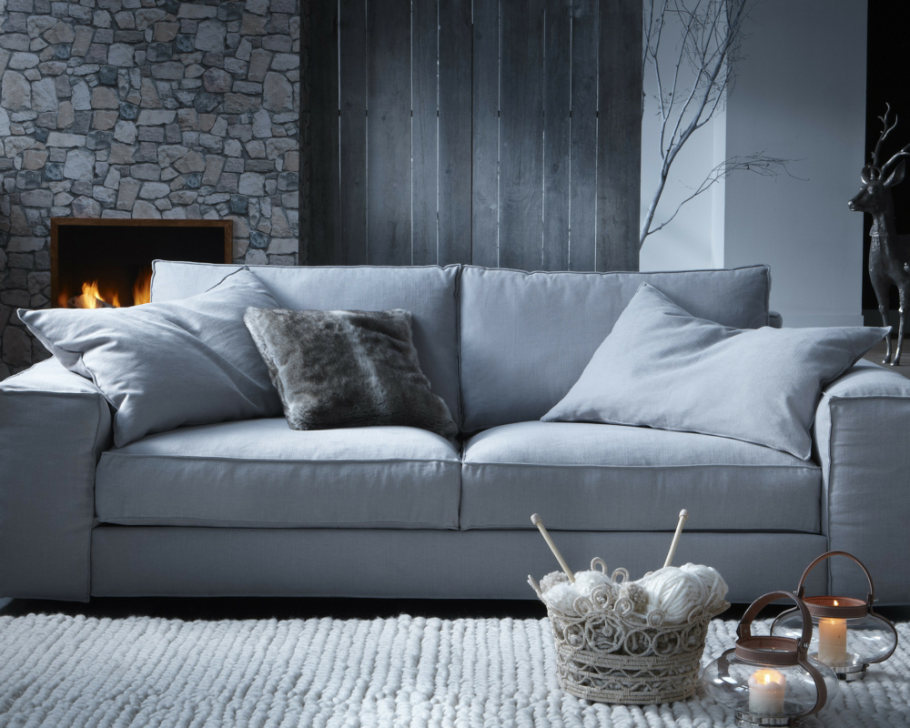 Canapé gris avec des coussins douillets dans un salon éclairé par des bougies et une cheminée, avec une décoration hivernale. Canape Paris by Atelier Bis