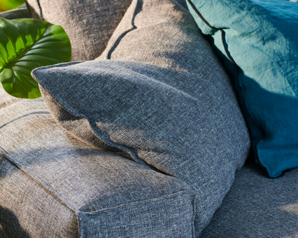 Gros plan sur un canapé en tissu gris avec un coussin bleu, à côté de plantes vertes en pot, en plein soleil. Canape Paris by Atelier Bis