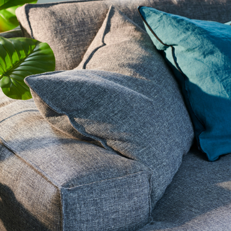 Gros plan sur un canapé en tissu gris avec un coussin bleu, à côté de plantes vertes en pot, en plein soleil. Canape Paris by Atelier Bis