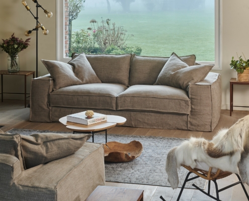 Salon moderne avec un canapé beige, un fauteuil, une table basse et une grande fenêtre donnant sur une cour verdoyante. Canape Paris by Atelier Bis