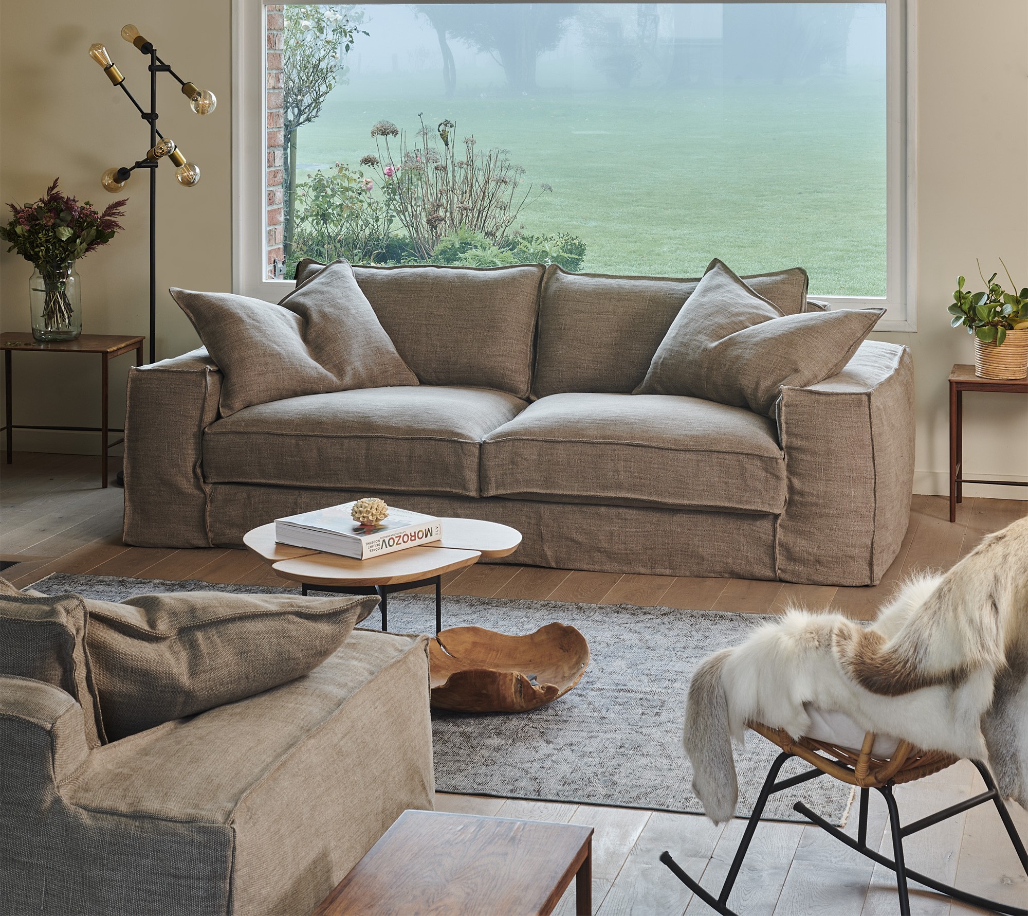 Salon moderne avec un canapé beige, un fauteuil, une table basse et une grande fenêtre donnant sur une cour verdoyante. Canape Paris by Atelier Bis