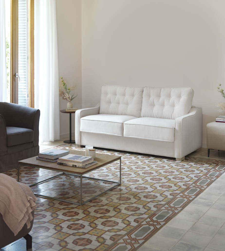 Salon lumineux avec un canapé convertible PAMPELUNE, un sol carrelé à motifs, une table basse et la lumière du soleil à travers des rideaux transparents. Canape Paris by Atelier Bis