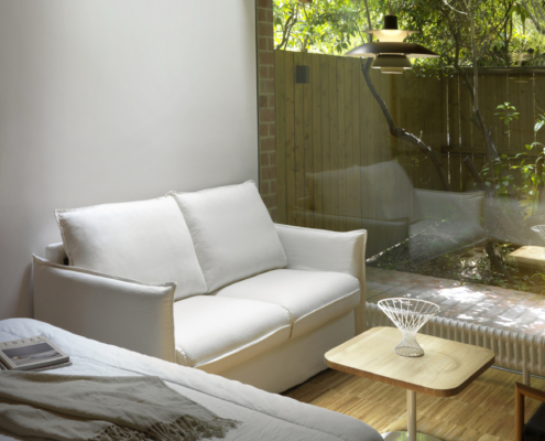 L'espace de vie minimaliste comprend un canapé convertible GRENADE, une table en bois, une grande fenêtre et une vue sereine sur le jardin à l'extérieur. Canape Paris by Atelier Bis