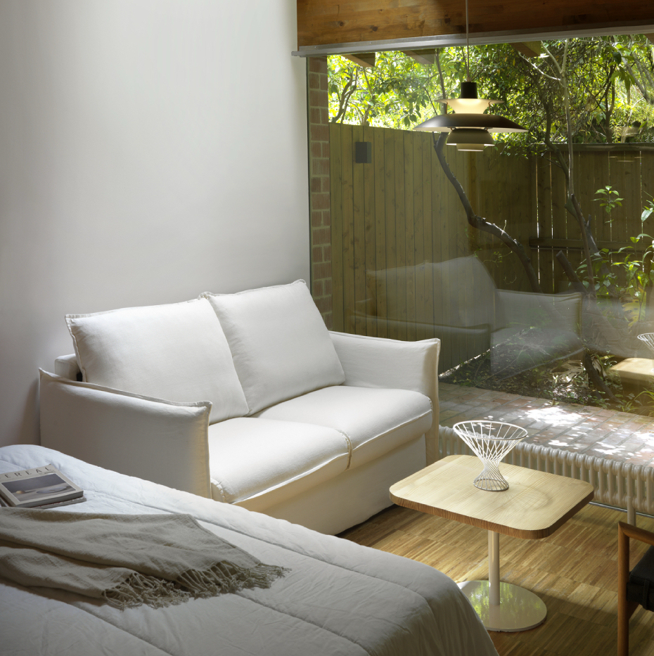 L'espace de vie minimaliste comprend un canapé convertible GRENADE, une table en bois, une grande fenêtre et une vue sereine sur le jardin à l'extérieur. Canape Paris by Atelier Bis