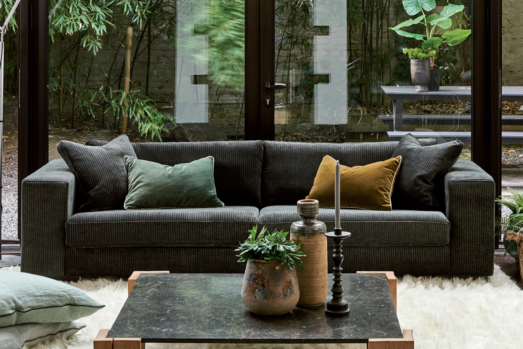 vang 2 Canapé foncé avec des coussins verts et dorés dans un salon confortable avec des plantes et une vue sur un jardin à travers des portes vitrées. Canape Paris by Atelier Bis