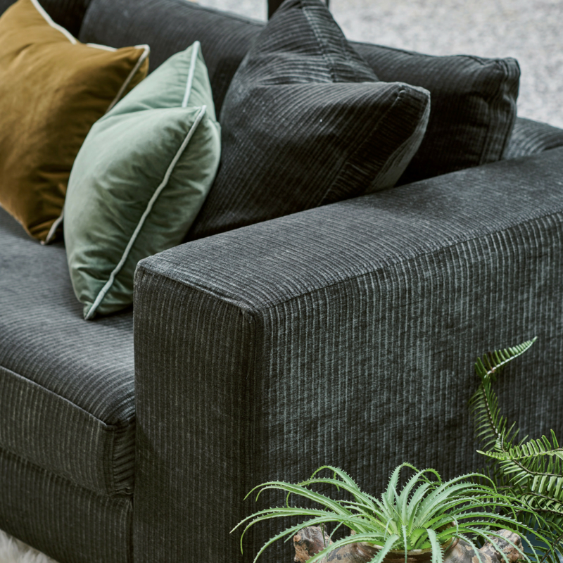 Canapé en velours côtelé gris foncé avec coussins verts et bruns, à côté d'une plante en pot sur un tapis blanc. Canape Paris by Atelier Bis