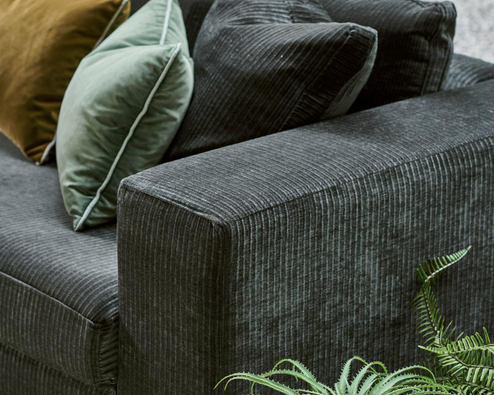 Canapé à la texture sombre avec des coussins verts et bruns, à côté d'une plante en pot et d'une grande fenêtre. Canape Paris by Atelier Bis